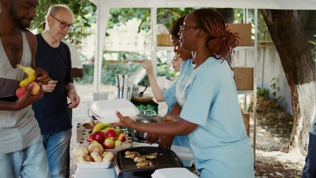 Generous volunteers wearing blue t-shirts, give free meals and humanitarian relief to those in need. Poor and homeless people of all races are fed by the non-profit organization. Side-view, handheld.