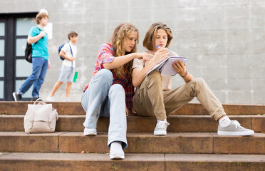 Fototapeta premium Two teenage students discussing while sitting with workbooks near college building on autumn day
