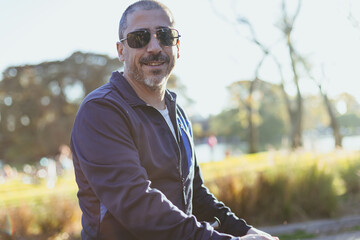 42-year-old man with gray hair, beard, sunglasses, smiling in a plaza