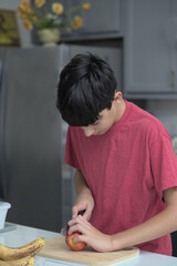 A boy cutting an apple with a knife in the kitchen counter wearing a red t-shirt.