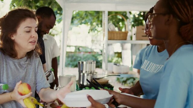 At outdoor food bank multiethnic female volunteers giving the homeless free food. Non-profit organization helps the hungry and underprivileged while providing humanitarian aid. Side-view handheld.