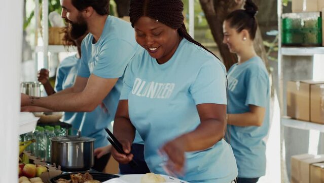 Black woman serving meals to underprivileged at community outreach event. Charity workers at a food drive distributing nutritious foods and essentials to the poor and homeless. Side-view, tripod shot.