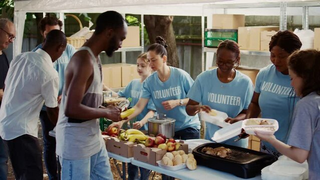 Multiethnic voluntary individuals distribute donated food, extending a helping hand to homeless and hungry people. Young volunteers share fresh, complimentary meals with those less fortunate.