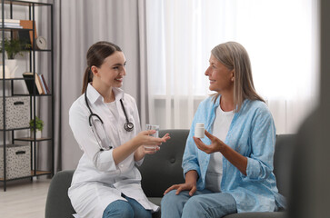 Young healthcare worker giving glass of water to senior woman with pills indoors