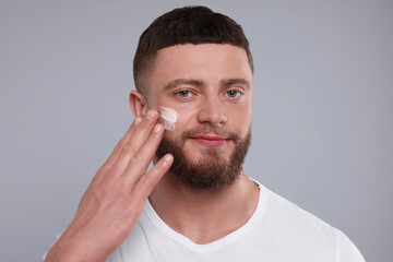 Handsome man applying cream onto his face on light grey background