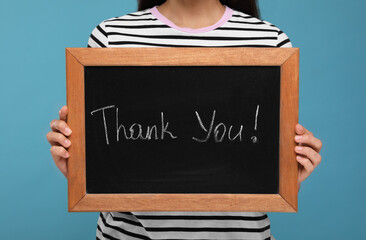 Woman holding small chalkboard with phrase Thank You on light blue background, closeup