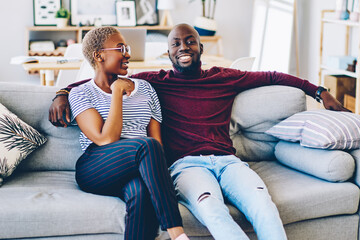 Obraz premium Beautiful dark skinned woman talking to smiling boyfriend sitting together on cozy sofa in living room,