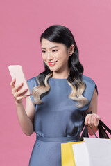 Confident fashionable young Asian woman in sexy dress holding gold shopping bags using mobile application on smartphone