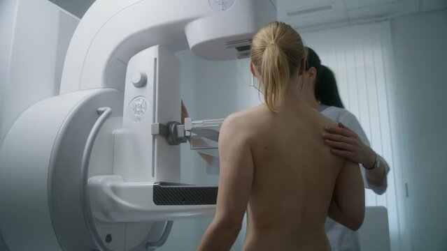 Hospital radiology room. Adult woman stands topless preparing for mammography screening procedure. Female doctor adjusts mammogram machine for patient. Breast cancer prevention. Modern bright clinic.