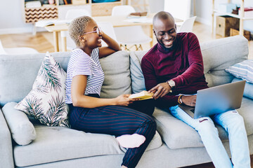 Cheerful african marriage having fun during free time at home sitting on cozy couch in living room
