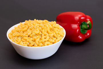 Uncooked Chifferi Rigati Pasta in White Cup and Red Bell Pepper on Black Background. Fat and Unhealthy Food. Scattered Classic Dry Macaroni. Italian Culture and Cuisine. Raw Pasta