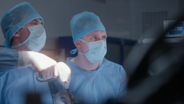 Face close up of two surgeons in lab coats using laparoscope during surgery. Doctors, nurses and paramedics perform surgical operating with seriously injured patient. Medical staff in modern clinic.