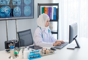 Asian professional successful experienced muslim female doctor radiologist in white lab coat with stethoscope sitting in radiography department with monitor screen office typing working with computer - Powered by Adobe