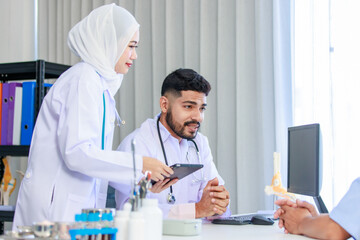 Asian Indian professional successful bearded male and muslim female doctors in white lab coat with stethoscope holding touchscreen tablet computer discussing with patient when making appointment