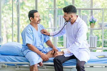 Obraz premium Asian Indian professional experienced young handsome bearded male doctor practitioner in white lab coat clinical uniform using stethoscope listening old senior elderly male patient heartbeat on bed