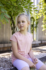 Portrait of a cute and pretty blonde little girl against a background of green broad-leaved plants....