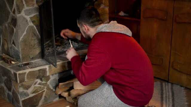 a young bearded man lights a fire in the fireplace in a hunting lodge. Cozy winter holiday atmosphere
