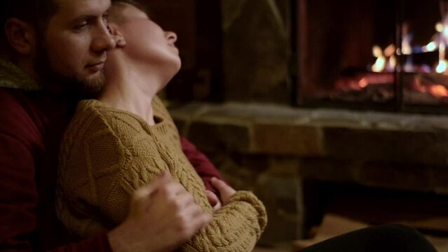 bearded man and handsome non binary girl with  short haircut and piercings are relaxing in the evening near the fireplace. Drinking tea in the cozy atmosphere of a hunting lodge, sitting near the fire