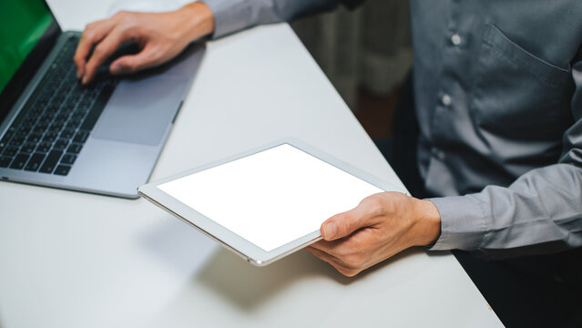Businessman top view hands holding white screen ipad touchpad mockup for website or application taking note using and typing on laptop showing media display information in corporate workplace table