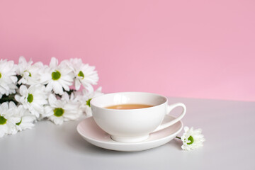 A white cup with tea and a saucer stands on a table with white chamomile flowers