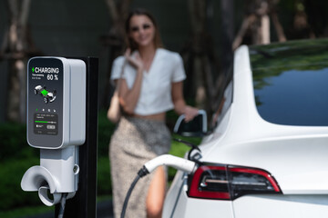 Young woman travel with EV electric car charging in green sustainable city outdoor garden in...