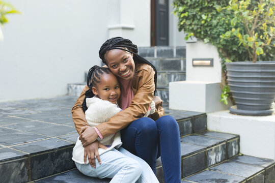 Happy african american mother and daughter embracing on steps in front of house - Powered by Adobe