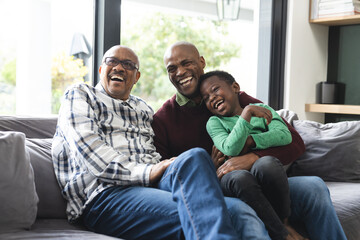 Happy african american father, son and grandfather sitting on couch,laughing,embracing, slow motion