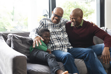 Happy african american father, son and grandfather sitting on couch,laughing and embracing