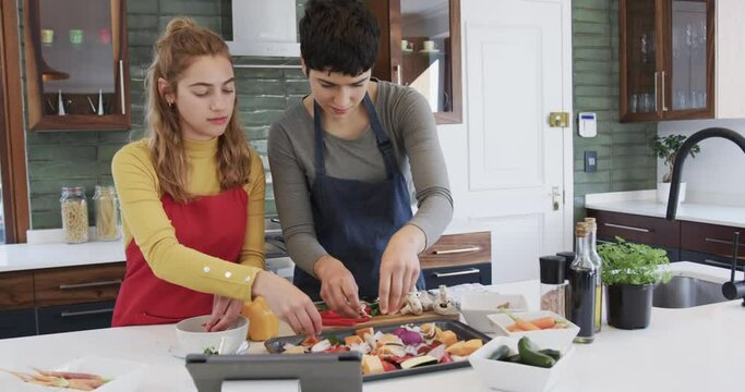 Happy caucasian lesbian couple preparing food and using tablet in sunny kitchen