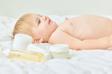 Obraz na płótnie Canvas Happy, lovely child, little baby girl lying on bed at home with skincare products. Moisturizing body with cream. Concept of childhood, kids cosmetology and natural cosmetics, body care