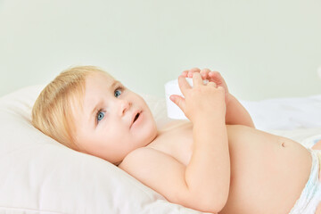 Cute toddler, little baby girl, child with blue eyes lying in bed and holding moisturizing body...