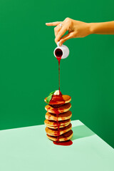 Food pop art photography. Close up. Female hand pouring sweet jam on delicious pancakes with butter over green background. Complementary colors.