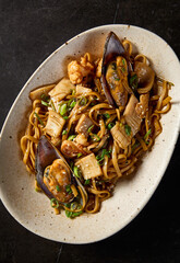 Close-up top view, capturing the intricate details of seafood-laden udon pasta, presented Asian-style, on a ceramic plate amidst a stark black setting