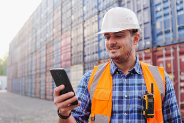 Young man engineer in the hard hat using phone in container warehouse. Logistics management in...