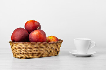 Red apples in a wicker basket and a cup on the wooden table.