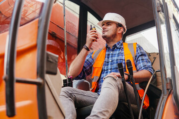 Young man loader worker or a forklift driver in a container warehouse wearing a white helmet is talking on the walkie-talkie or radio phone.