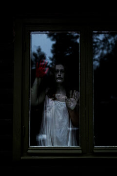 Female ghost with blood on hand seen through glass window of haunted cabin