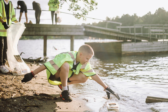 Full length of boy crouching while reaching for plastic at lakeshore