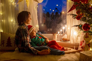 Toddler child, cute blond boy, sitting on the window in pajama, looking out for Satna Claus, Christmas lights around him