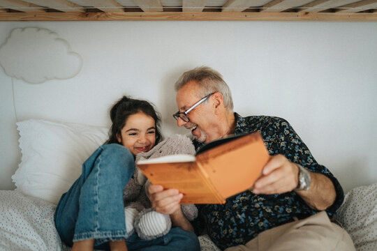 Happy senior man reading storybook with granddaughter on bed at home