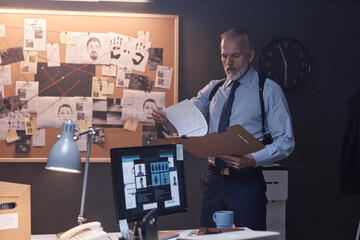 Waist up portrait of elegant senior detective reading file in office standing by evidence board,...