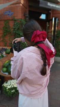 The European farmer's market features a charming young lady in pink, photographing the fresh produce.