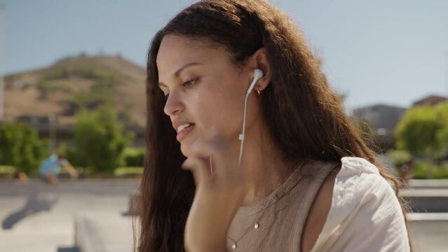 Young multiracial woman in casual clothing smiling while putting in headphones at the skatepark