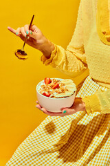 Woman in nice outfit holding golden spoon and bowl with muesli, cereal with berries against yellow background. Concept of healthy food, nutrition, pop art style, taste. Poster. Copy space for ad