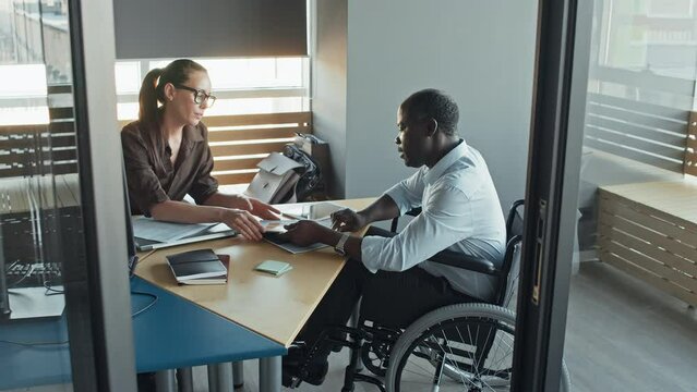 High angle zoom slowmo shot of black male employee in wheelchair talking to Caucasian female boss in office with business document