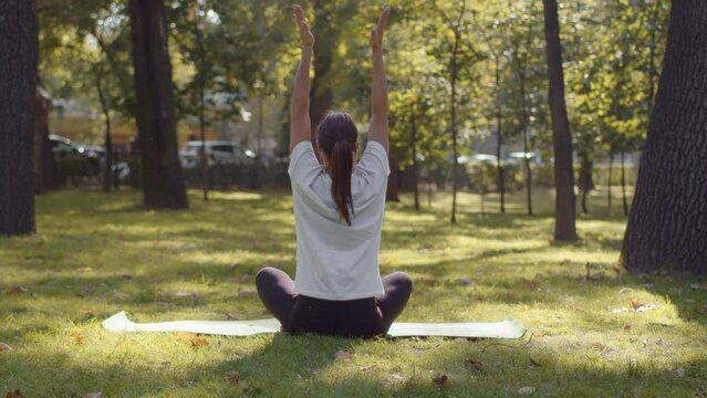 Young yoga woman sitting in the lotus pose on the yoga mat, raising her hands and folding them in a namaste gesture in the city park on a sunny day. Concept of a healthy, comfortable and conscious