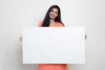 Indian woman in ethnic wear holding white sign board in hand and looking towards the camera ,...