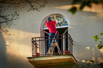 Man on balcony painting facade. Man stands on folding mini ladder and paint window jamb, renovating...