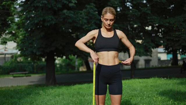 Front view of muscular woman wearing black sport uniform standing on open area while measuring amount of slim waist. Sportive caucasian female athlete monitoring girth of body regularly outdoors.