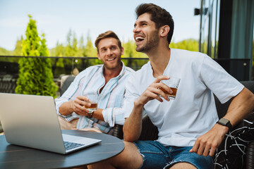 Two male friends drinking whiskey and using laptop while relaxing on terrace in mountains